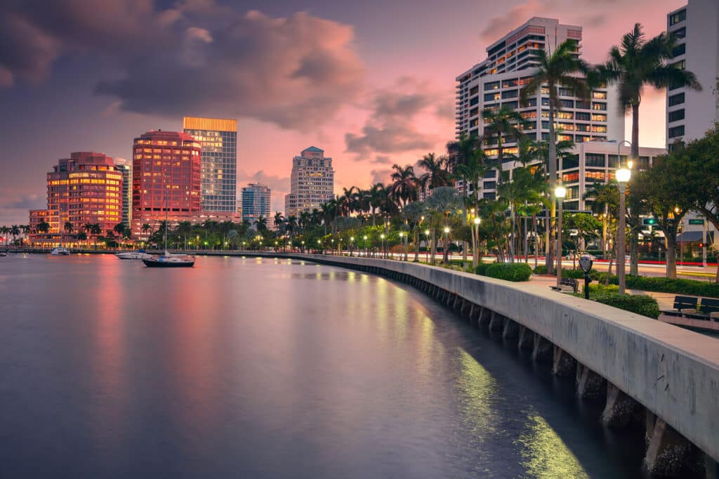 Die beleuchtete Uferpromenade in West Palm Beach, Florida, bei Sonnenuntergang. Palmen und Straßenlaternen säumen den Gehweg entlang des Wassers, das die Lichter der Stadt und den rosafarbenen Himmel widerspiegelt. Im Hintergrund stehen moderne Hochhäuser.