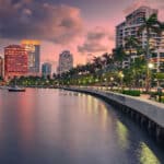 Die beleuchtete Uferpromenade in West Palm Beach, Florida, bei Sonnenuntergang. Palmen und Straßenlaternen säumen den Gehweg entlang des Wassers, das die Lichter der Stadt und den rosafarbenen Himmel widerspiegelt. Im Hintergrund stehen moderne Hochhäuser.