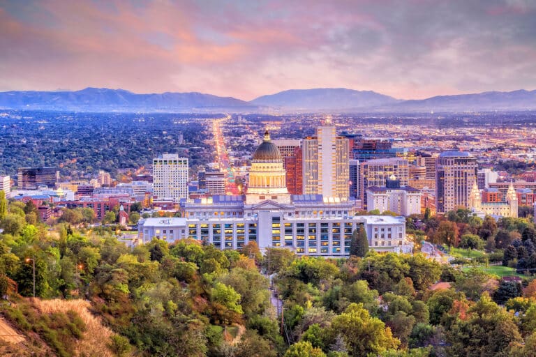 Abendstimmung in Salt Lake City Das Bild zeigt die Skyline von Salt Lake City im Abendlicht. Im Vordergrund steht das markante Utah State Capitol mit seiner großen Kuppel, umgeben von Bäumen und Grünflächen. Dahinter erstreckt sich die moderne Innenstadt mit zahlreichen Hochhäusern und Gebäuden, die sich bis zu den Ausläufern der Rocky Mountains erstreckt. Der Himmel ist in weichen Rosa- und Lilatönen gefärbt, was der Szene eine stimmungsvolle Atmosphäre verleiht.