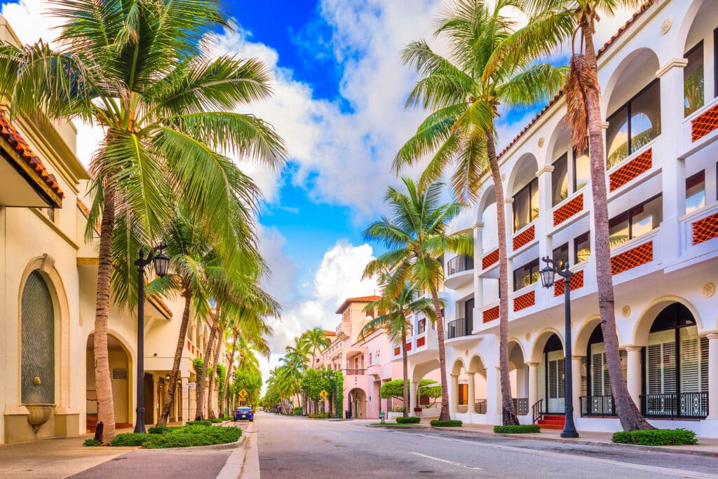 Dieses Bild zeigt eine Allee in Boca Raton, Florida, die von Palmen und Gebäuden im spanischen Stil gesäumt ist. Der Himmel ist blau mit weißen Wolken.