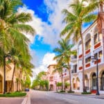 Dieses Bild zeigt eine Allee in Boca Raton, Florida, die von Palmen und Gebäuden im spanischen Stil gesäumt ist. Der Himmel ist blau mit weißen Wolken.