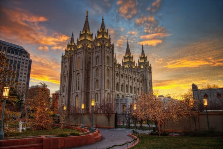 Der Salt-Lake-Tempel in Salt Lake City bei Sonnenuntergang. Das große, gotische Gebäude ist hell beleuchtet und steht vor einem dramatischen Himmel mit orangefarbenen und grauen Wolken. Die Bäume im Vordergrund sind mit Lichtern geschmückt.
