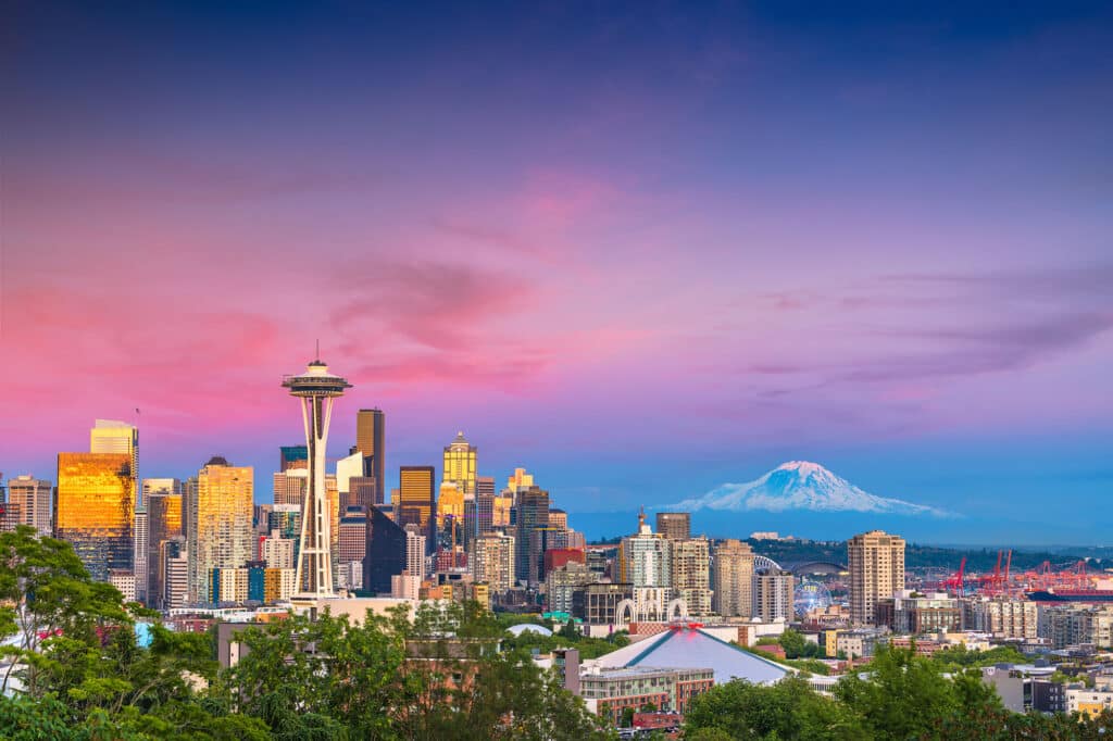Die Skyline von Seattle bei Sonnenuntergang. Im Vordergrund sind Bäume zu sehen, dahinter erstrecken sich die Wolkenkratzer der Stadt, mit der markanten Space Needle im Zentrum. Im Hintergrund erhebt sich der schneebedeckte Mount Rainier. Der Himmel ist in leuchtenden Violett- und Rosatönen gefärbt.