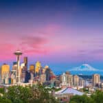 Die Skyline von Seattle bei Sonnenuntergang. Im Vordergrund sind Bäume zu sehen, dahinter erstrecken sich die Wolkenkratzer der Stadt, mit der markanten Space Needle im Zentrum. Im Hintergrund erhebt sich der schneebedeckte Mount Rainier. Der Himmel ist in leuchtenden Violett- und Rosatönen gefärbt.
