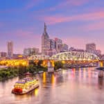 Die beleuchtete Skyline von Nashville, Tennessee, bei Sonnenuntergang. Ein Raddampfer-Schiff fährt auf dem Cumberland River im Vordergrund. Die Shelby Street Pedestrian Bridge führt über den Fluss und verbindet das Stadtzentrum mit den östlichen Stadtteilen.