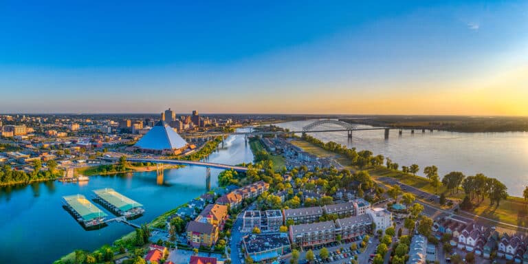 Ein breites Panorama von Memphis, Tennessee, bei Sonnenuntergang. Die Aufnahme zeigt die Skyline der Stadt am Ufer des Mississippi River, die die große, pyramidenförmige Bass Pro Shops at the Pyramid und die Hernando de Soto Bridge umfasst. Die untergehende Sonne taucht den Himmel und das Wasser in ein warmes, goldenes Licht.
