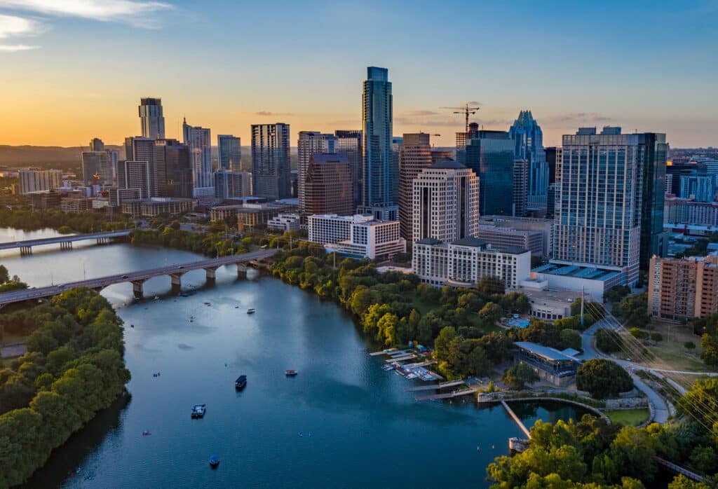 Die Skyline von Austin, Texas, bei Sonnenuntergang. Im Vordergrund fließt der Colorado River, auf dem sich einige Boote befinden. Die Stadt mit ihren modernen Wolkenkratzern erstreckt sich am Flussufer. Der Himmel ist in sanften Orange- und Blautönen gefärbt.
