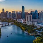 Die Skyline von Austin, Texas, bei Sonnenuntergang. Im Vordergrund fließt der Colorado River, auf dem sich einige Boote befinden. Die Stadt mit ihren modernen Wolkenkratzern erstreckt sich am Flussufer. Der Himmel ist in sanften Orange- und Blautönen gefärbt.