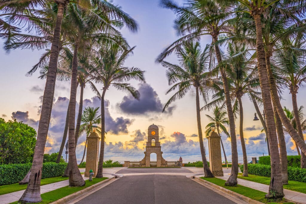 Die Allee zum Strand in Boca Raton, Florida, bei Sonnenuntergang. Die Straße ist von hohen Palmen gesäumt, die zum ikonischen Uhrenturm am Meer führen. Der Himmel ist in sanften Lila-, Orange- und Rosatönen gefärbt.