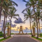 Die Allee zum Strand in Boca Raton, Florida, bei Sonnenuntergang. Die Straße ist von hohen Palmen gesäumt, die zum ikonischen Uhrenturm am Meer führen. Der Himmel ist in sanften Lila-, Orange- und Rosatönen gefärbt.