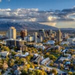 Die Skyline von Salt Lake City, Utah, bei Sonnenuntergang. Die Stadt liegt in einem Tal, umgeben von schneebedeckten Bergen. Die Bäume im Vordergrund zeigen Herbstfarben.