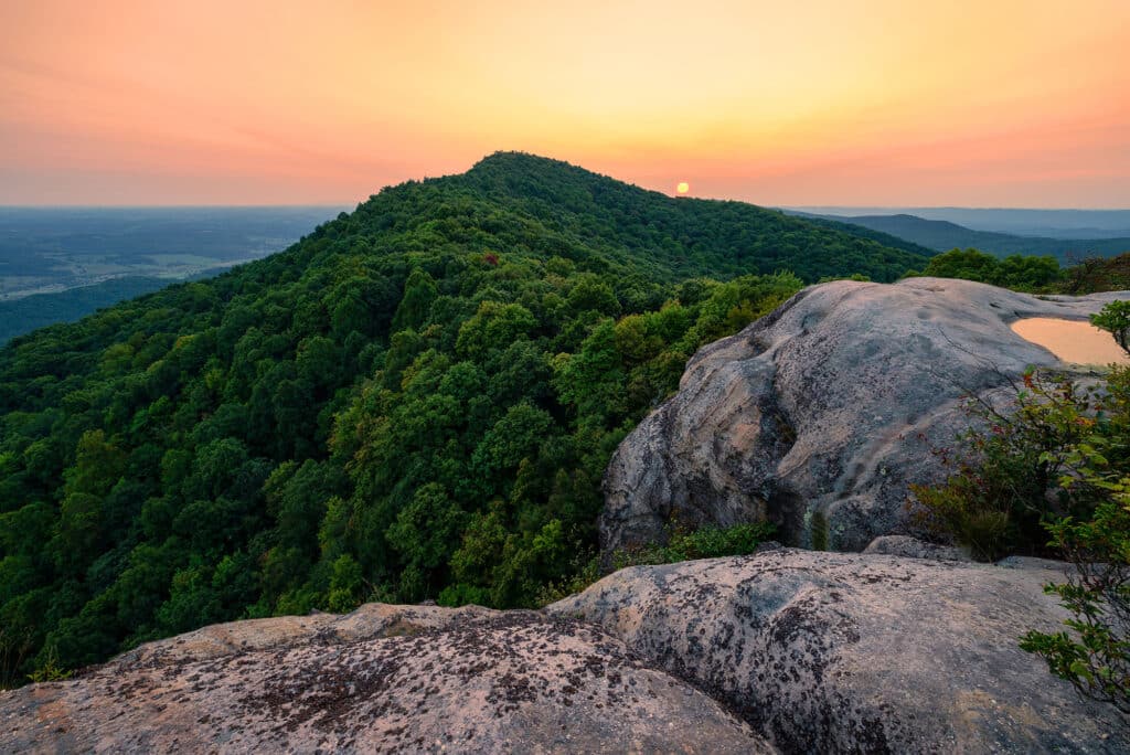 Ein Panoramablick von White Rock im Cumberland Gap Nationalpark. Die Aufnahme zeigt einen bewaldeten Hügel und eine weite Landschaft unter einem Himmel bei Sonnenauf- oder Sonnenuntergang, der in einem warmen, orangen Licht leuchtet. Im Vordergrund sind große, helle Felsen zu sehen.