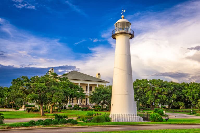 Ein großer weißer Leuchtturm in Biloxi, Mississippi, mit einem historischen Haus und grünen Bäumen im Hintergrund bei Sonnenuntergang.