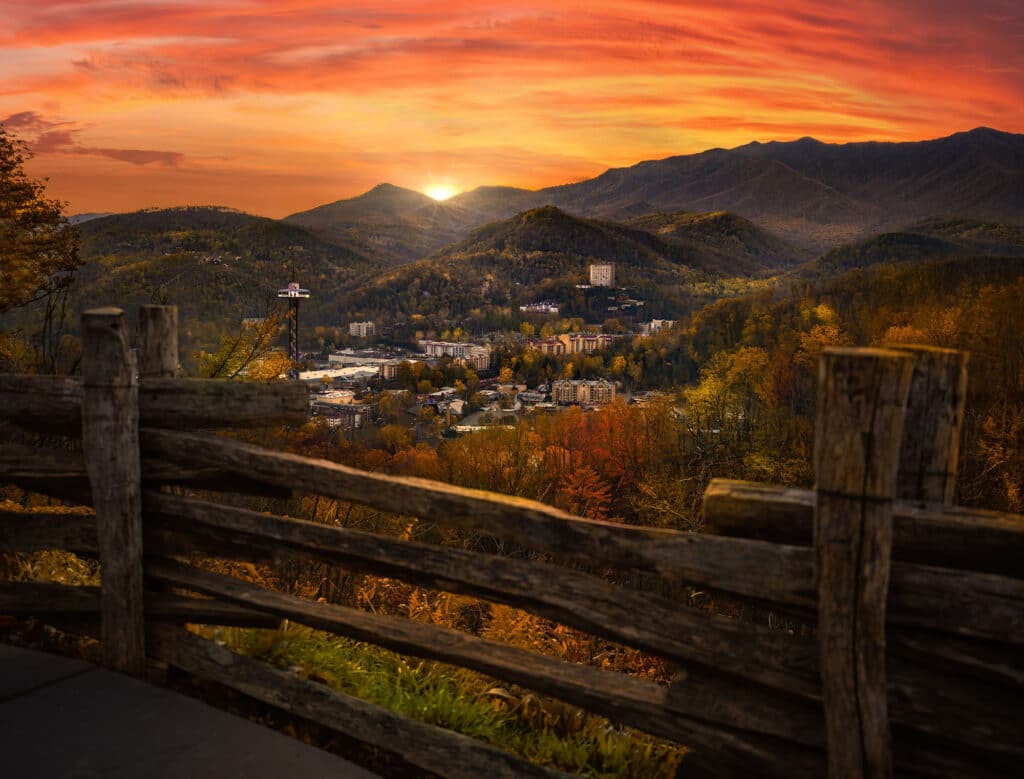 Landschaftsaufnahme der Stadt Gatlinburg, Tennessee, und der umliegenden Berge bei Sonnenuntergang. Im Vordergrund ist ein Holzzaun zu sehen.