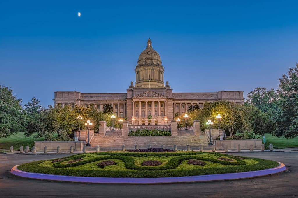 Das Kentucky State Capitol in Frankfort bei Dämmerung, beleuchtete Laternen und ein gepflegter Vorplatz sind im Vordergrund. Der Mond ist am Himmel sichtbar.